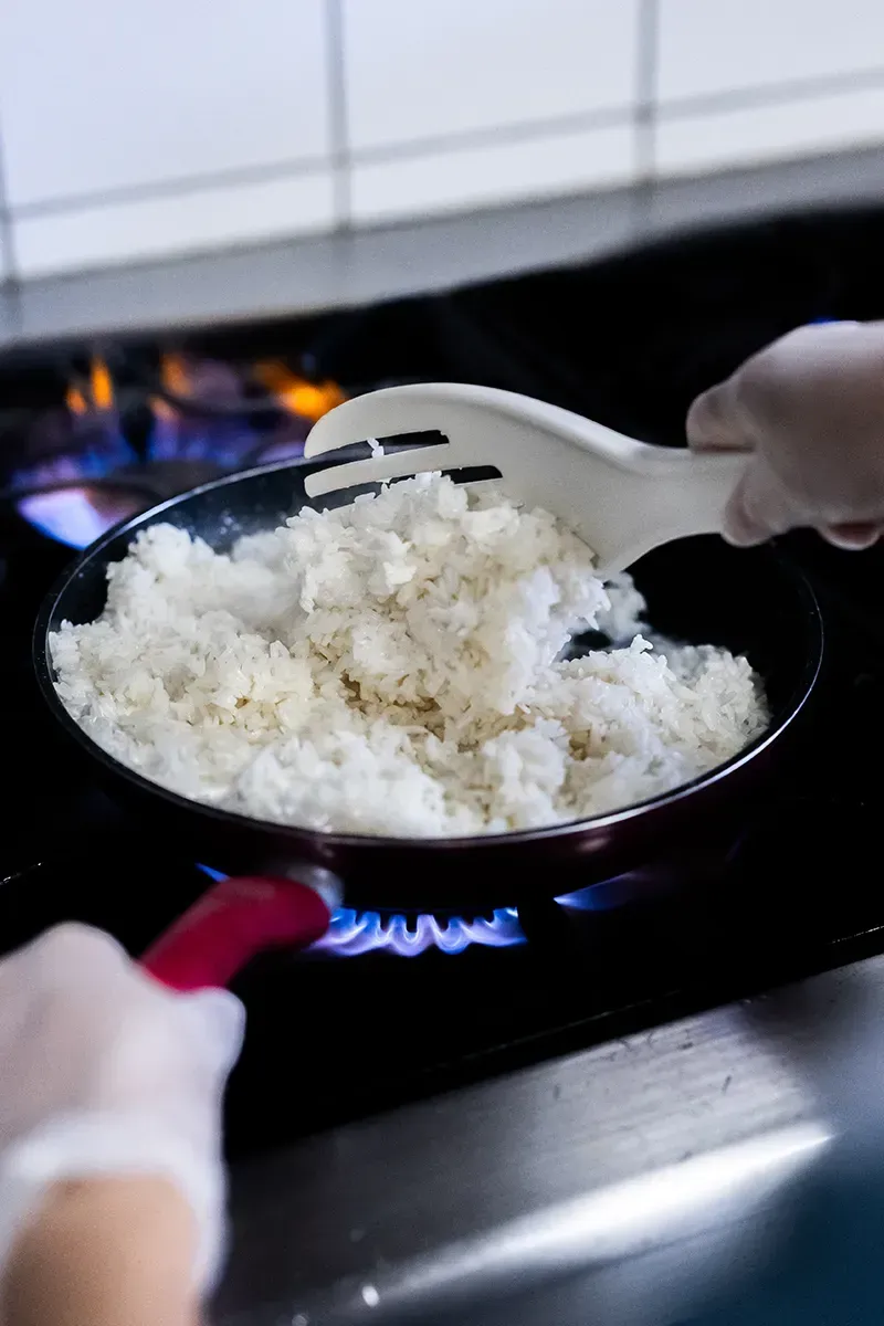 Une personne remue du riz blanc dans une poêle sur une flamme de gaz bleue à l'aide d'une spatule blanche.