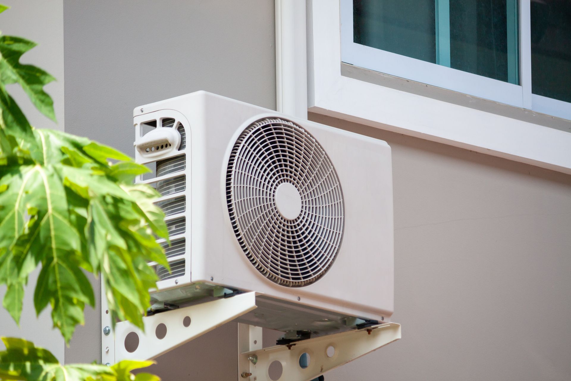White air conditioning unit mounted on a gray wall, next to a window and green leaves. White air conditioning unit mounted on a gray wall, next to a window and green leaves.