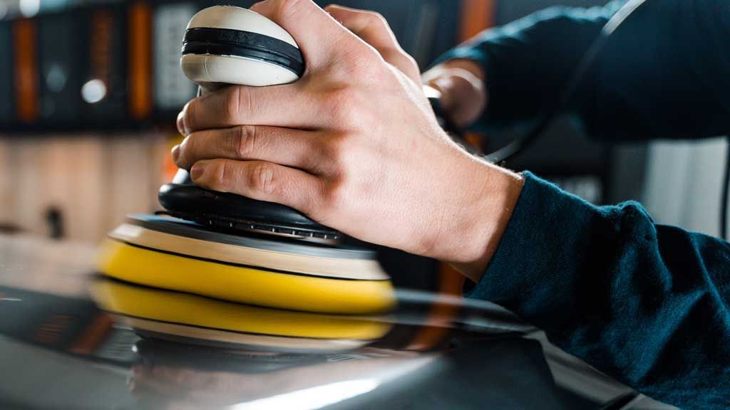 Person polishing a car's surface with an electric buffer; hand close-up, yellow buffing pad.