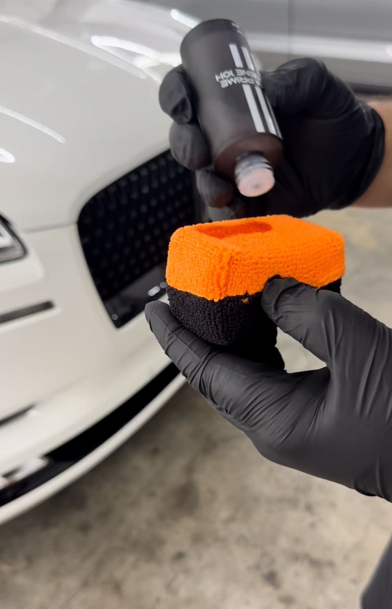 Gloved hands applying ceramic coating from a black bottle onto an orange applicator pad, on a white car.