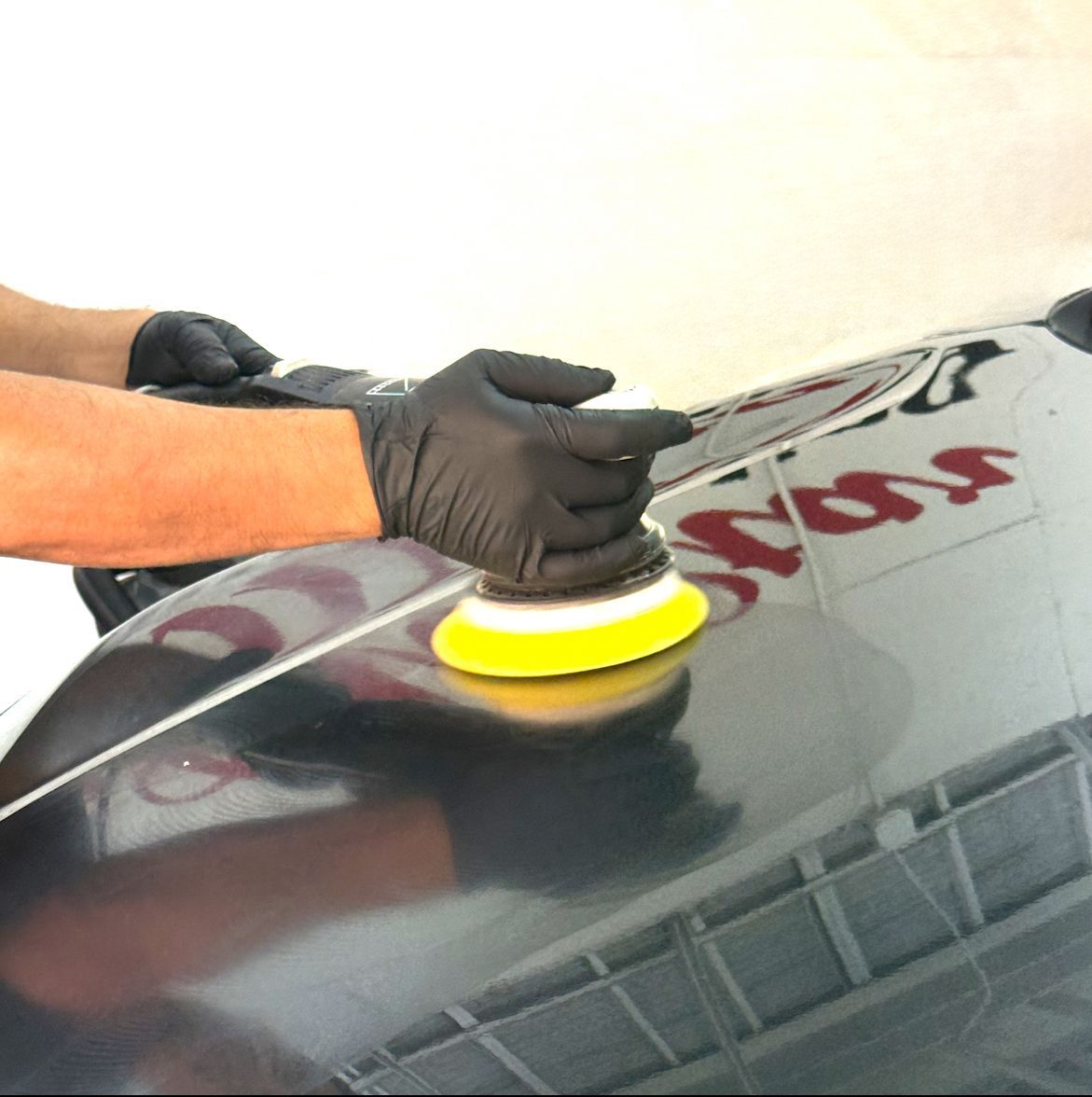 Person in black gloves polishing a black car with a yellow pad.