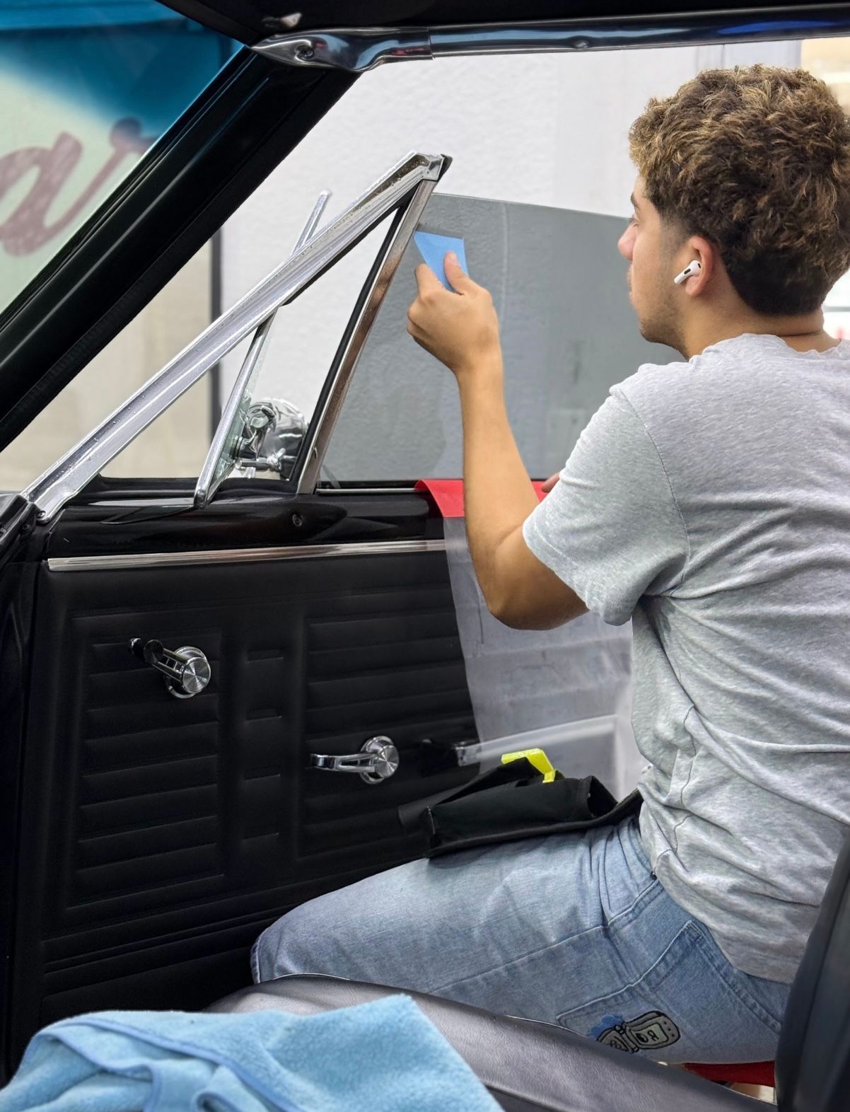 A man installing window tint in a classic car, using a blue tool. Inside a garage, he wears a gray shirt and jeans.