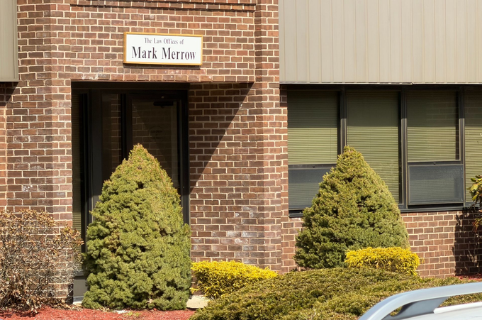 Brick building entrance with sign above door, windows, and shrubs in front.