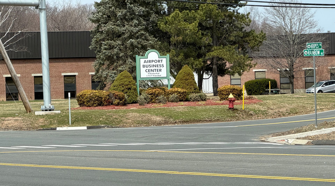 Street view of a brick building with a white sign, shrubs, and trees beside a road.