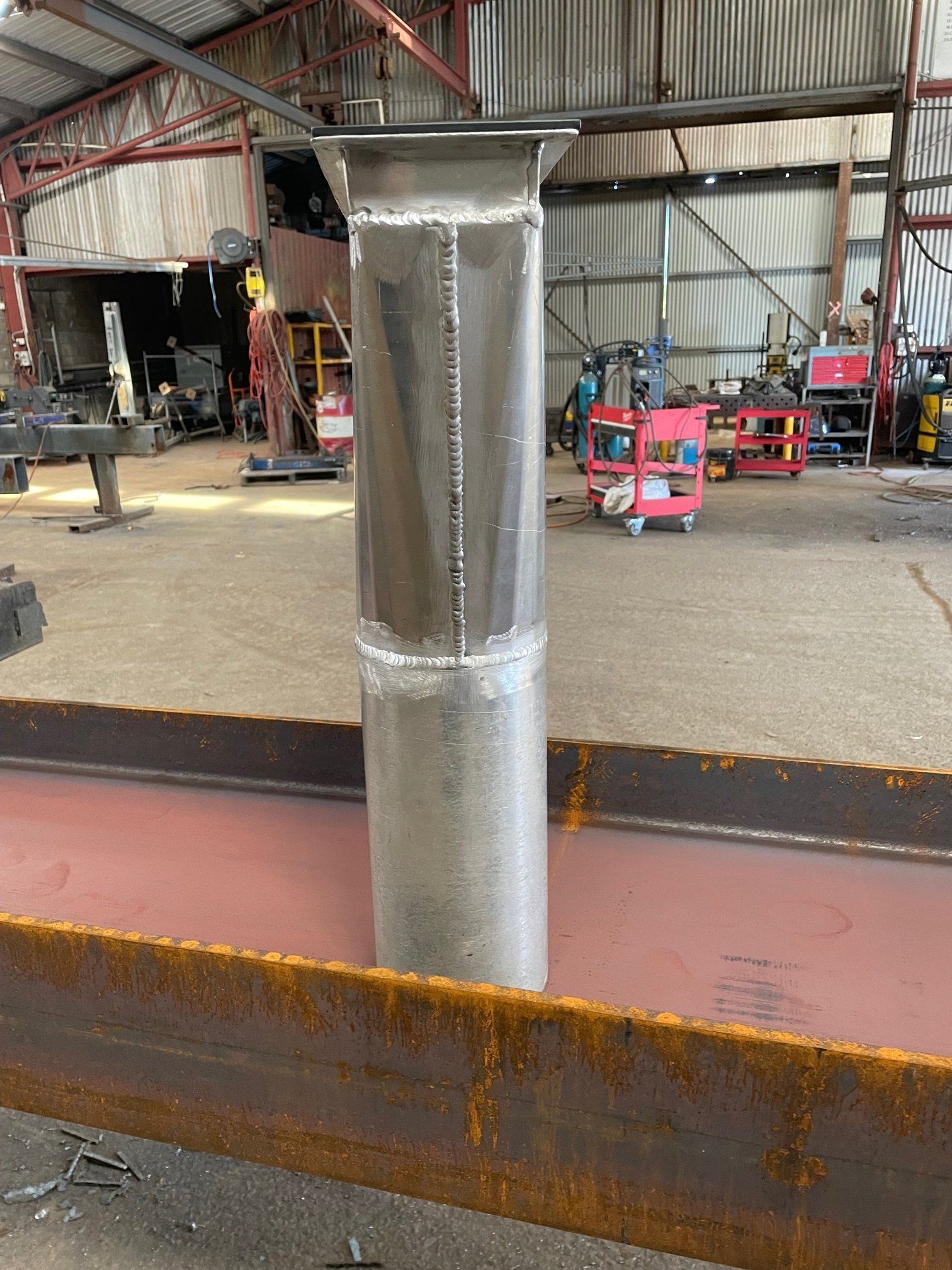 A stainless steel pipe is sitting on top of a rusty metal beam in a factory.