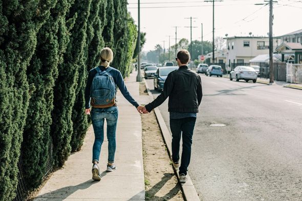 Couple walking hand-in-hand down a sidewalk along a road, next to a tall green hedge.