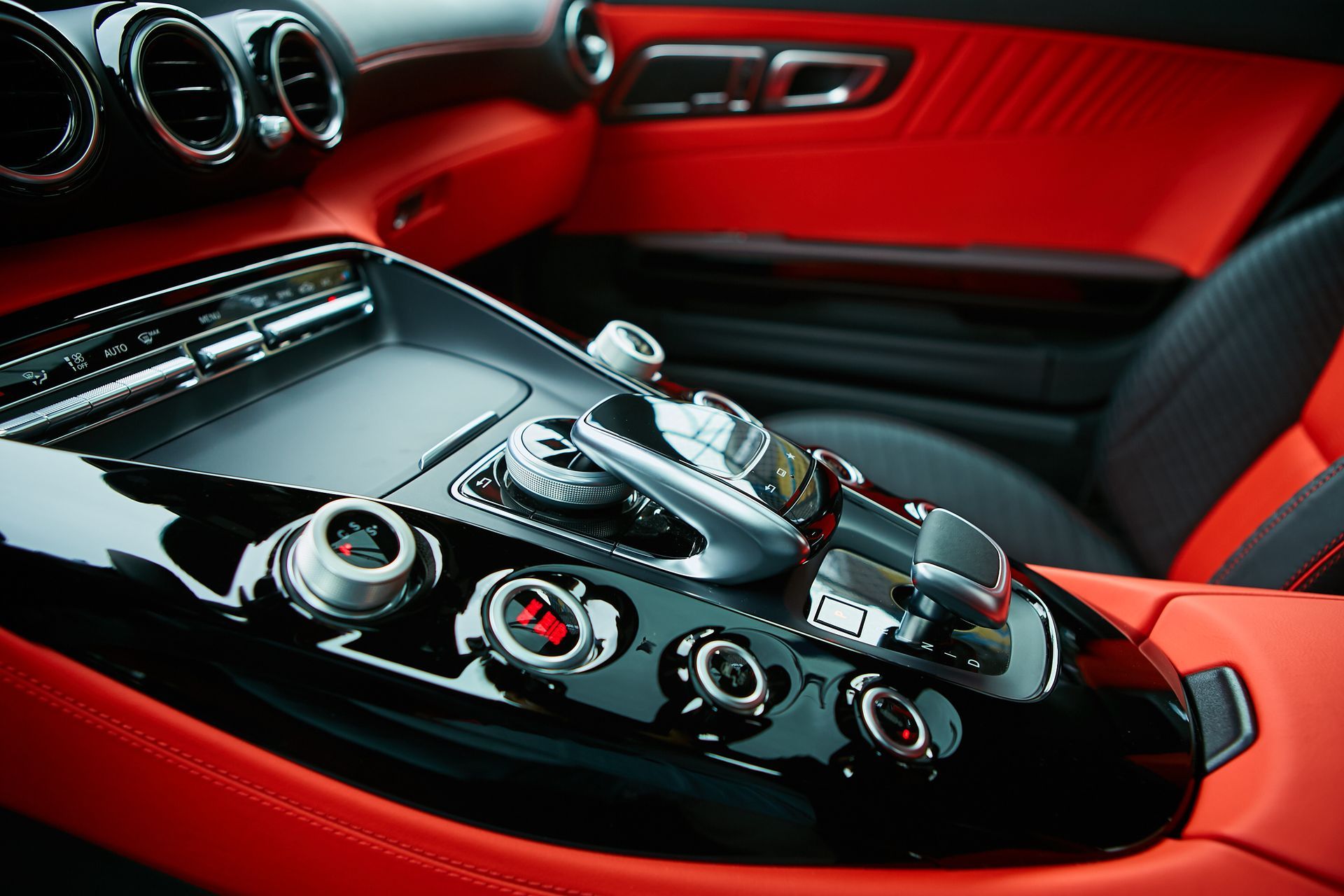 Close-up of a luxury car interior. Black glossy console with silver controls, surrounded by red leather.