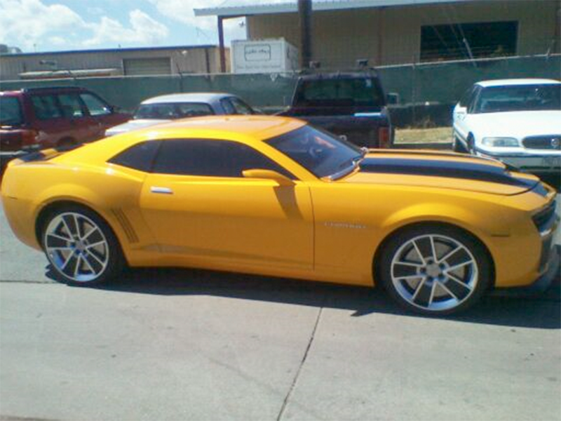 A bright yellow Chevrolet Camaro with black racing stripes parked in a sunny outdoor lot.