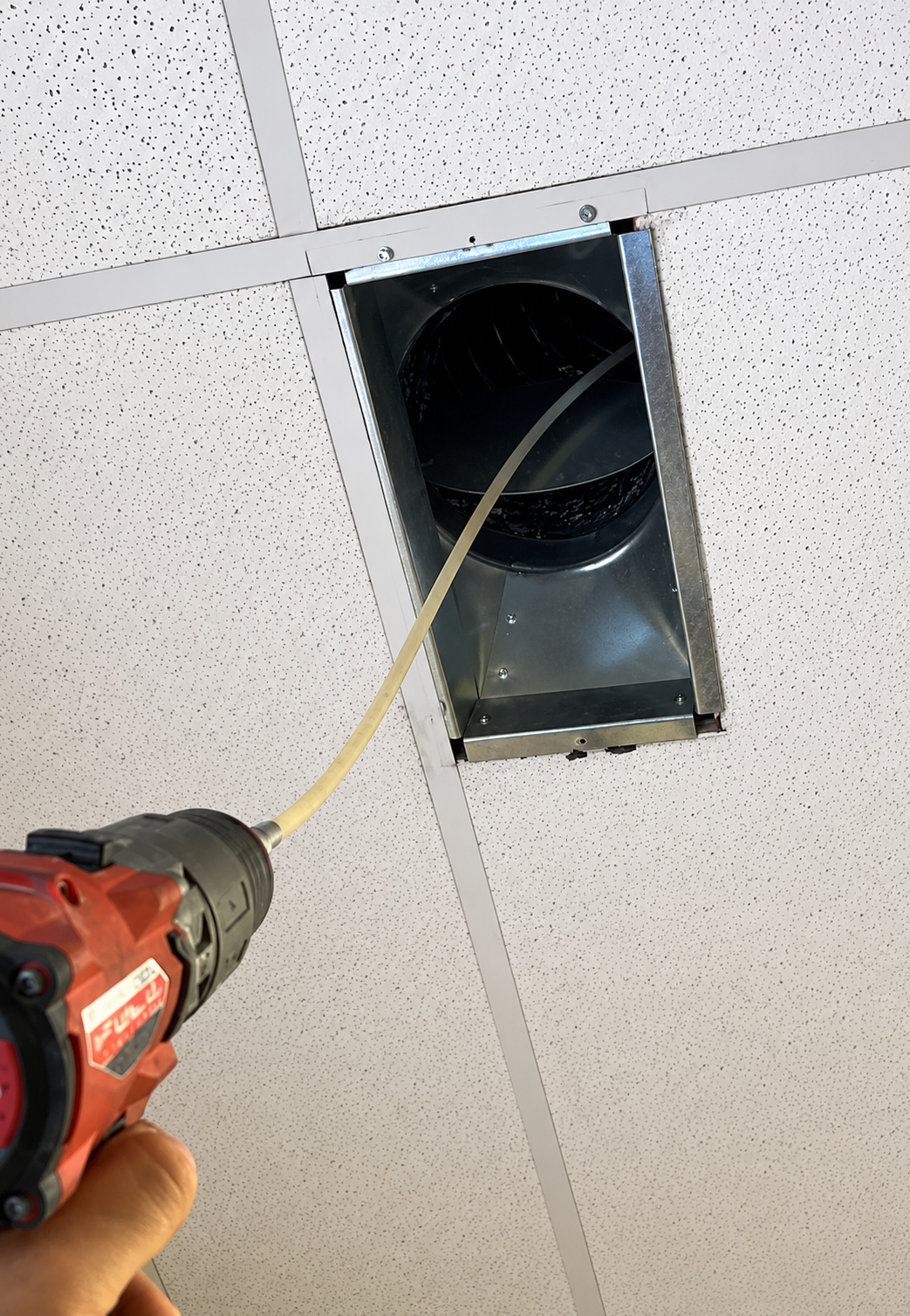 A person drills a ceiling tile beside a vent opening while a cable is fed through the hole.
