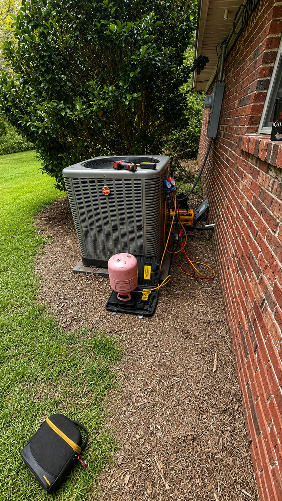 Outdoor HVAC unit beside a brick house, with tools and a pink refrigerant tank on gravel
