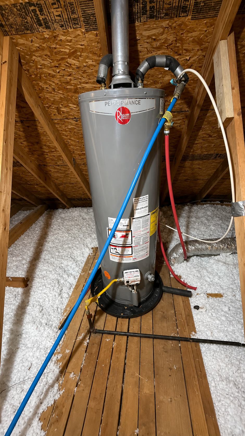 Water heater in an attic with blue and red hoses attached, standing on a wooden floor beneath exposed insulation.