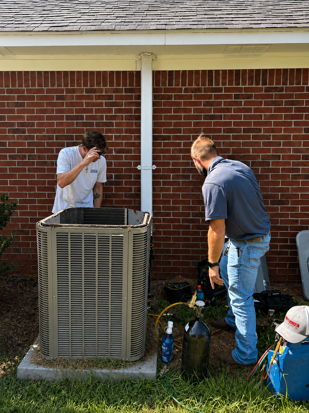 Two men working on an outdoor air conditioner unit beside a brick house.