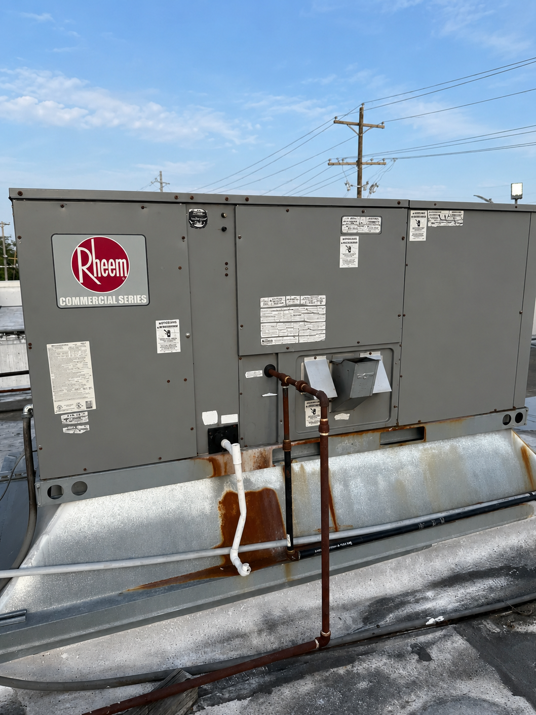 Gray rooftop HVAC unit with red logo and rusted piping under a blue sky