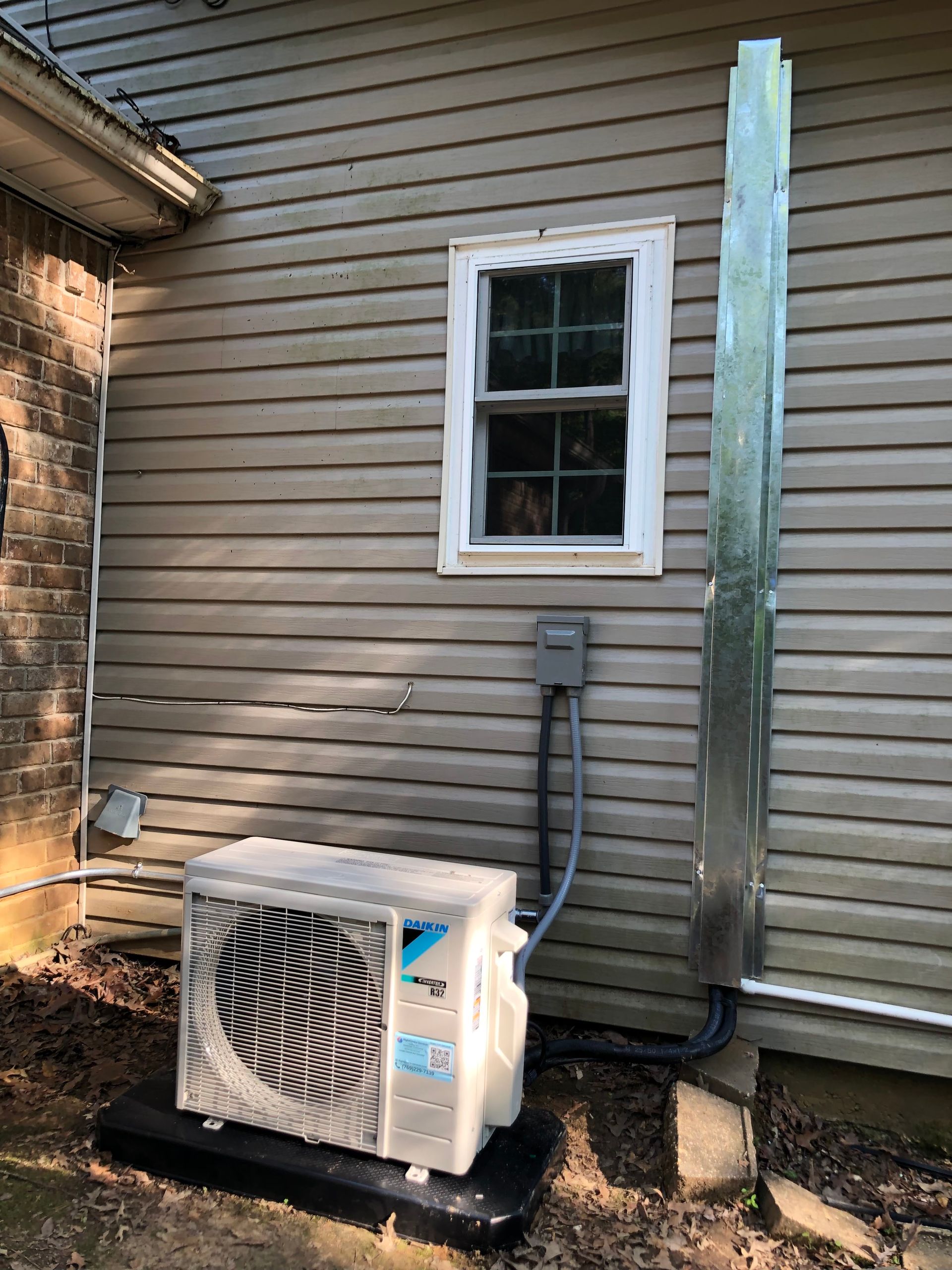 Outdoor HVAC unit beside a beige house wall under a small window, with exposed utility piping