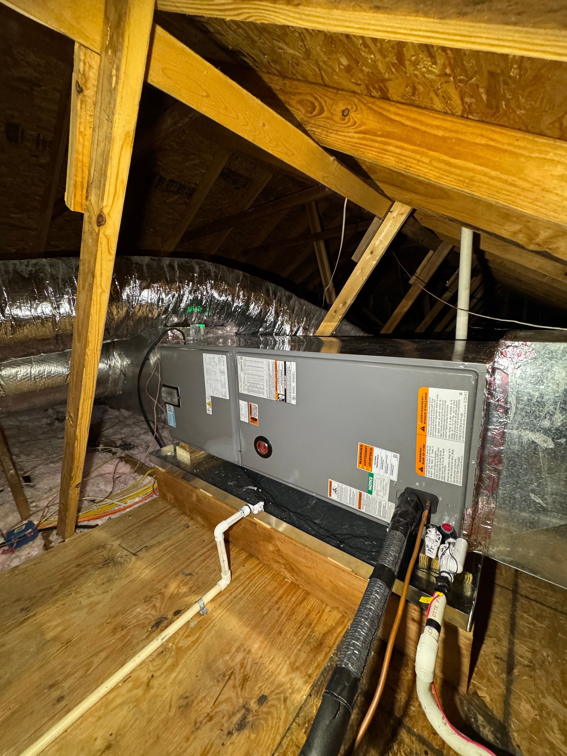 Attic HVAC unit amid insulation and exposed wooden beams, with ductwork and wiring visible.