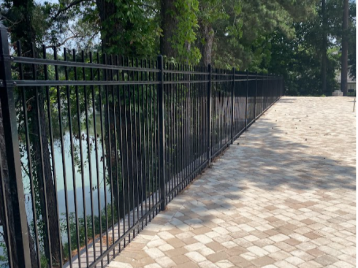 Black metal fence beside a paved path, with trees and water on the left in a park-like setting