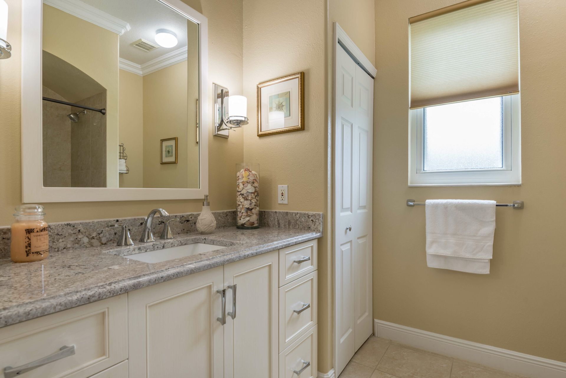 Bright beige bathroom with granite vanity, mirror, towel rack, and window blinds.