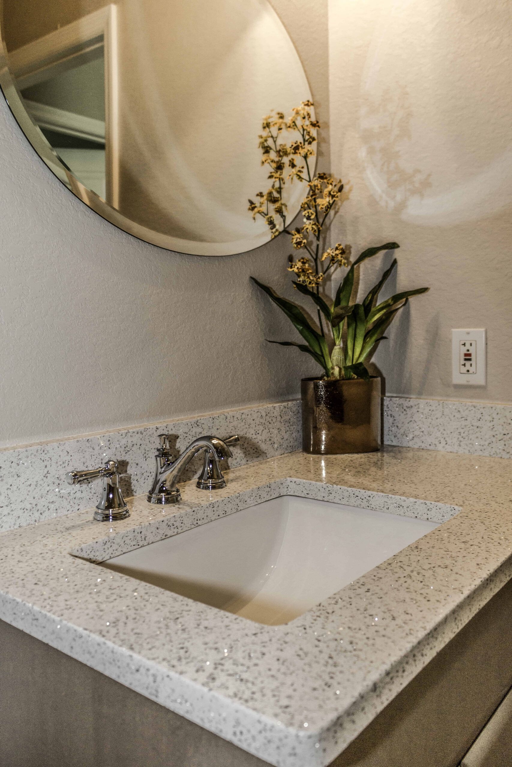 Modern bathroom vanity with speckled countertop, white sink, chrome faucet, round mirror, and orchid vase