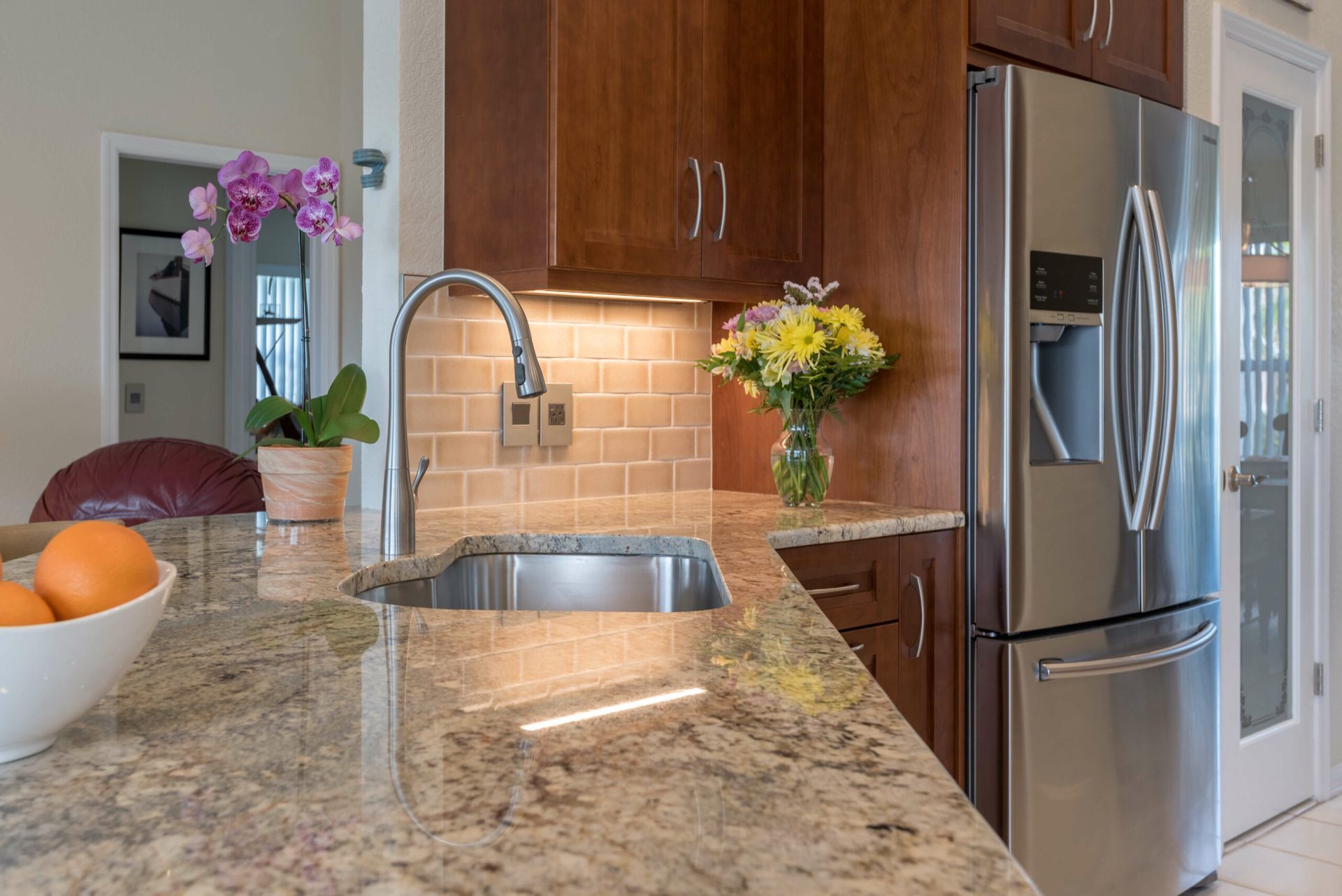 Modern kitchen with granite countertop, sink, stainless fridge, and flowers by the window.