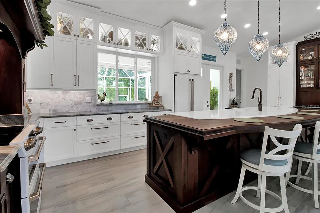 Bright white kitchen with dark island, pendant lights, large window, and a white chair by the counter