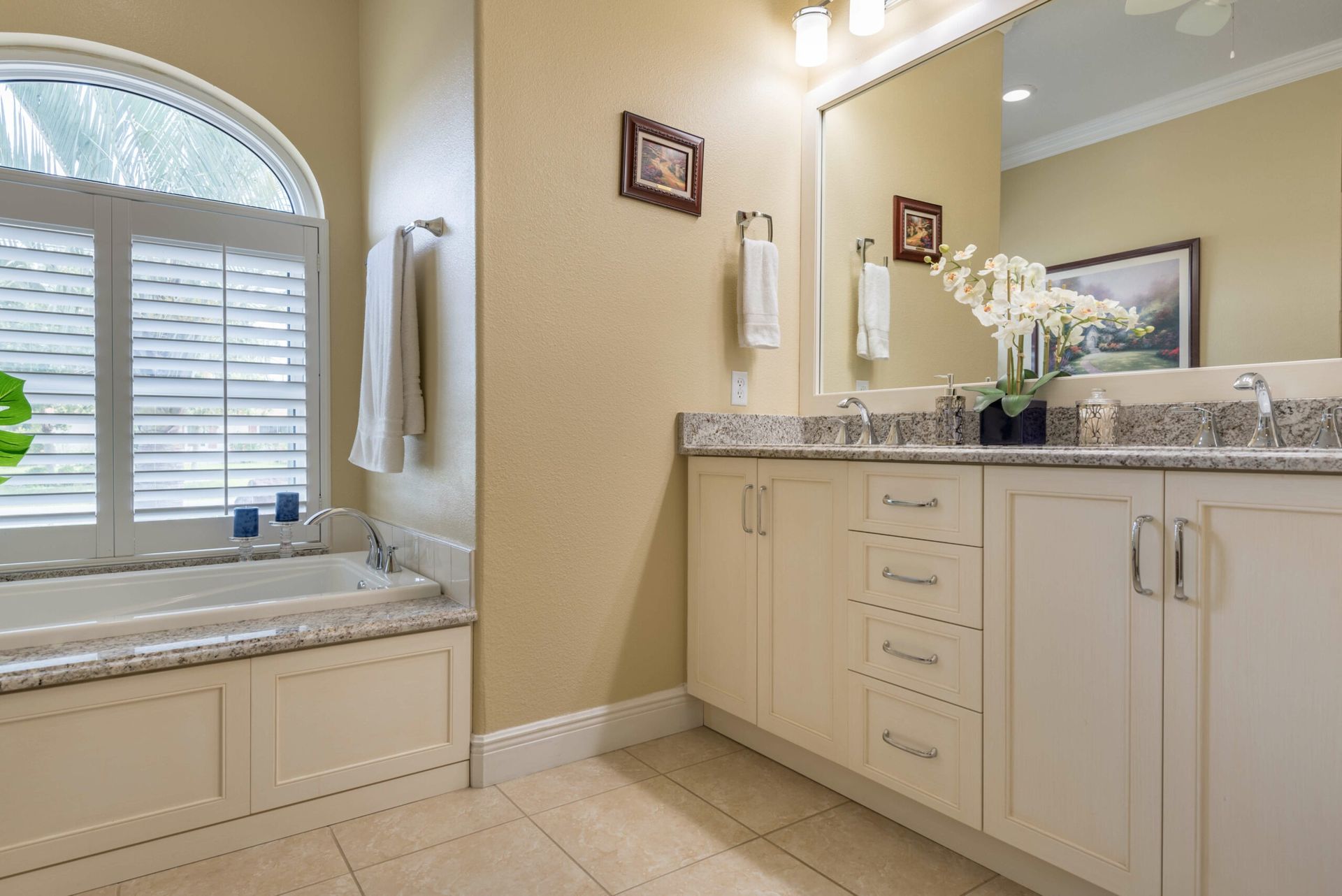 Bright bathroom with a double vanity, large mirror, tiled floor, and window above a built-in bathtub.