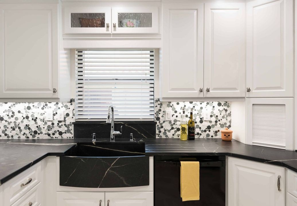 Modern white kitchen with black countertops, sink, window blinds, and mosaic backsplash
