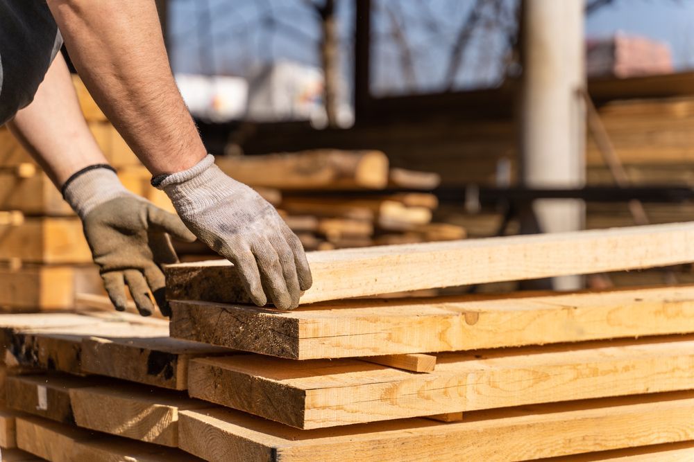 A Man Wearing Gloves is Stacking Wooden Boards on Top of Each Other — TM Built Construction In Lochinvar, NSW
