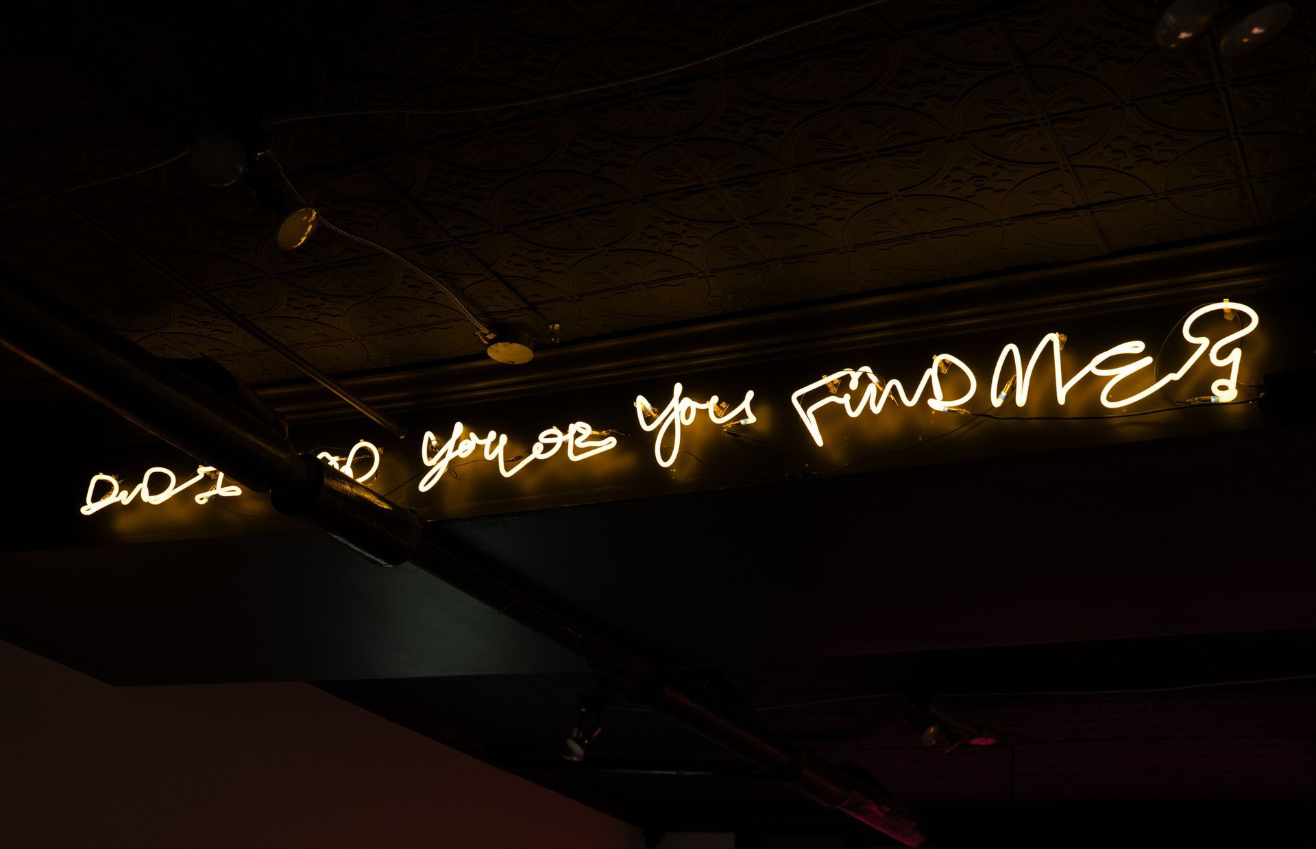 Yellow neon sign on ceiling: "Does no one find me?" ; art by Yale Wolf.