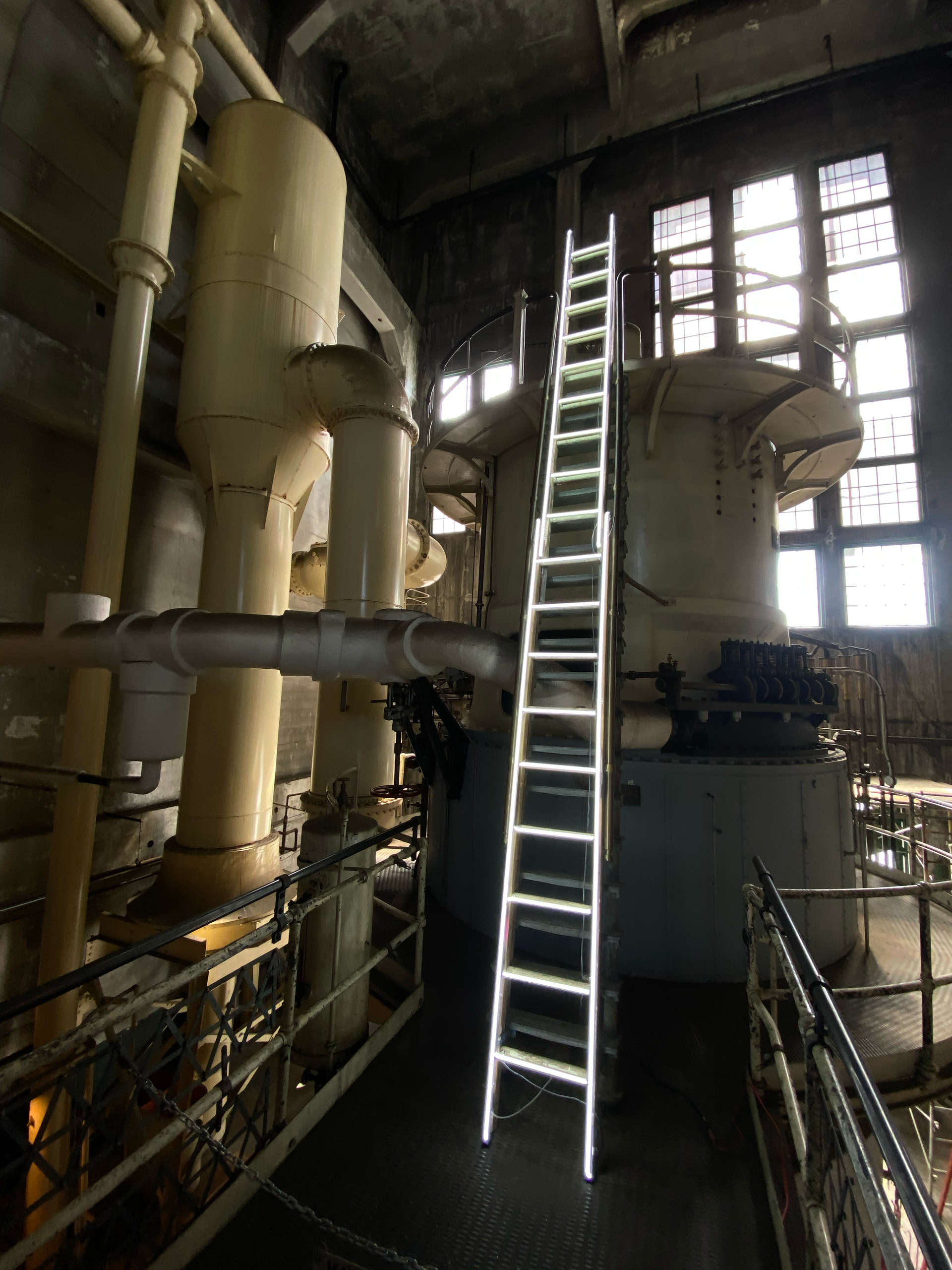 Tall ladder leans against large industrial machinery in a dimly lit, high-ceilinged room; art by Yale Wolf.