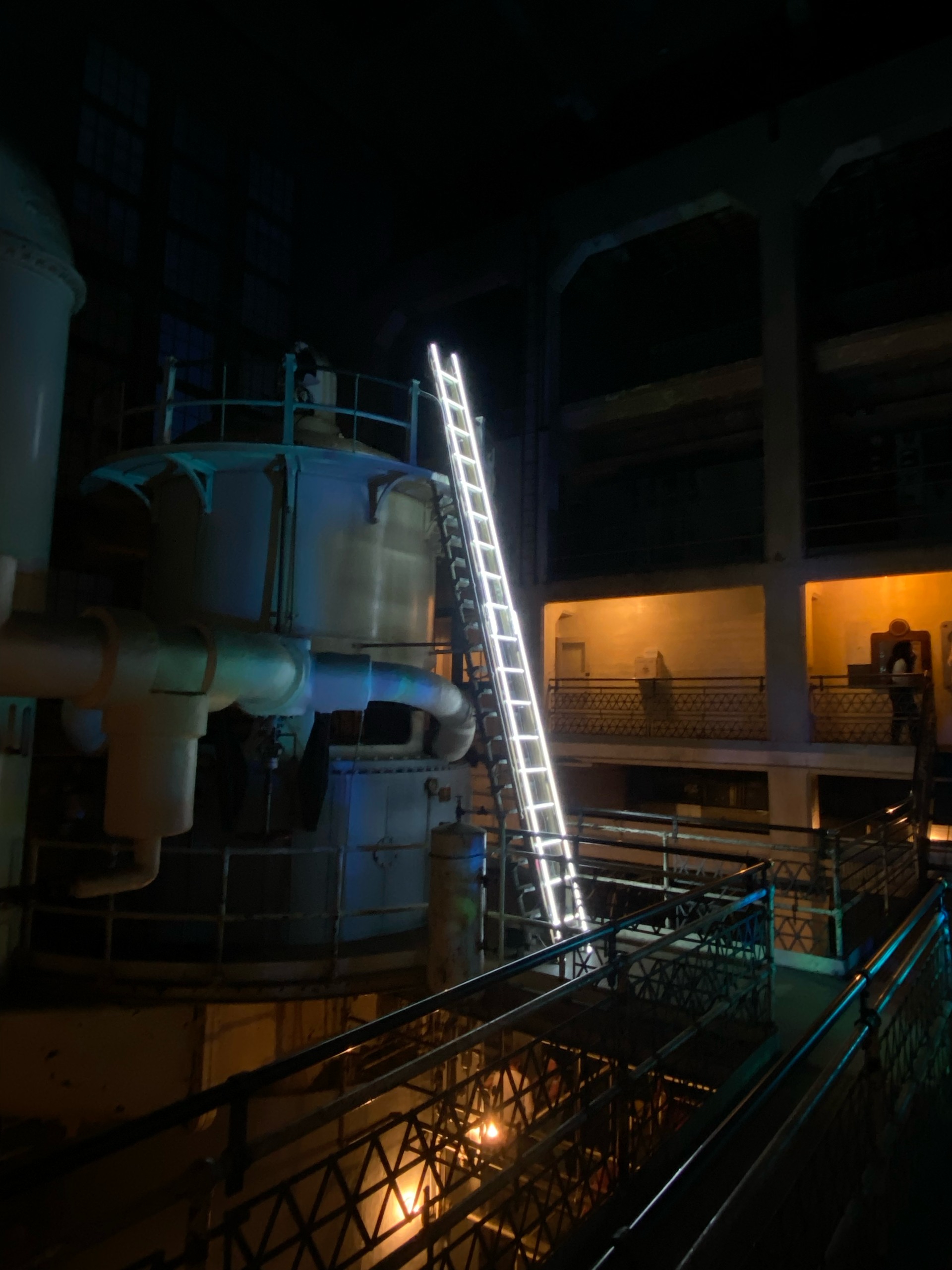 Ladder lit against large industrial equipment in dimly lit space; art by Yale Wolf. 