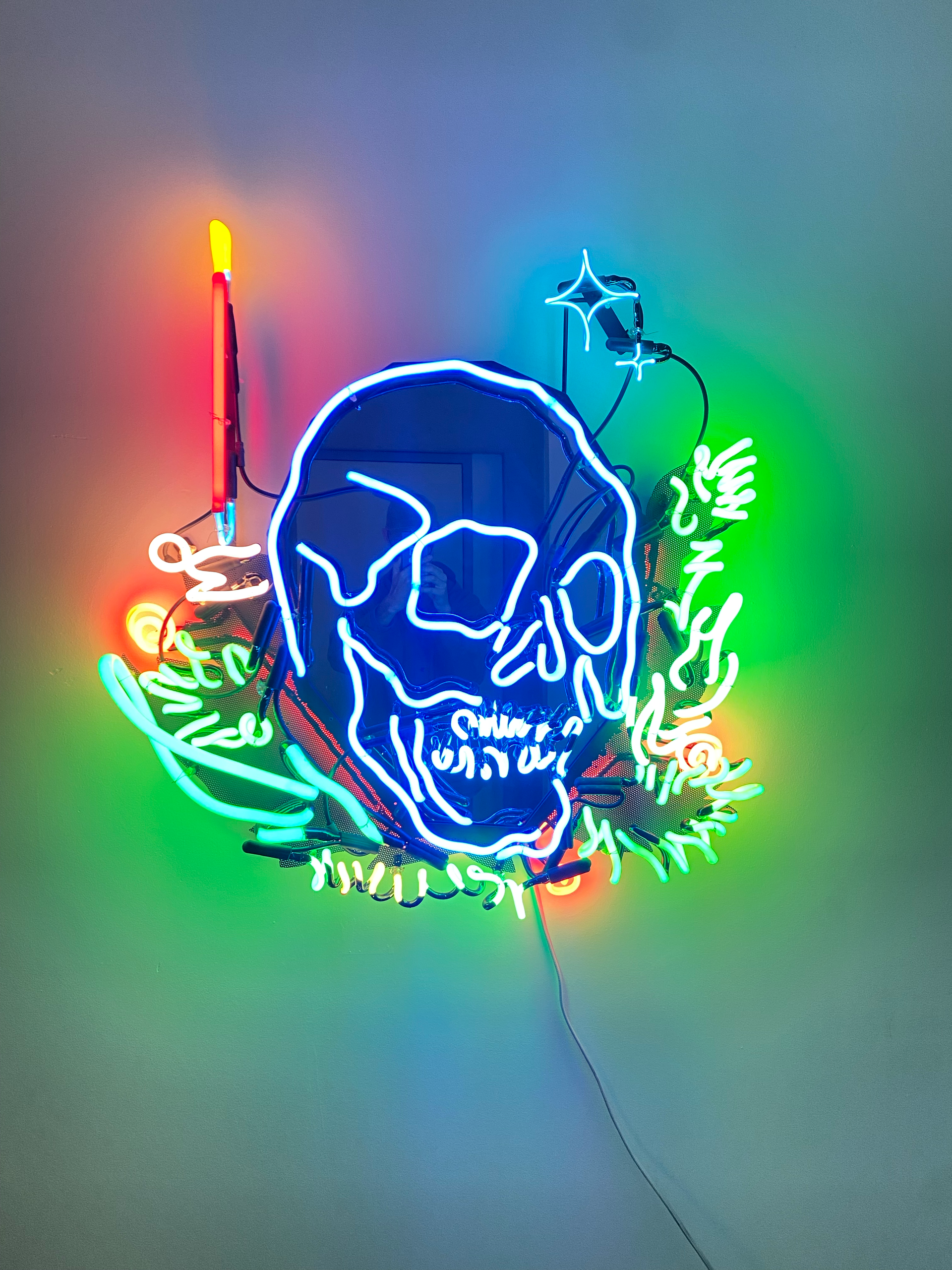 Neon skull, candle, and foliage in blues, greens, and reds on a wall; by Yale Wolf.