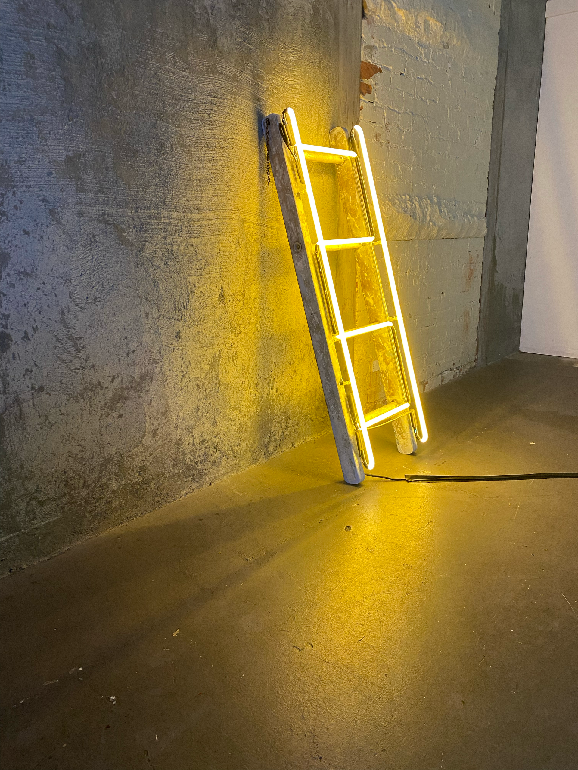 Yellow neon ladder by Yale Wolf leans against a rough gray wall, casting a bright shadow on the floor.