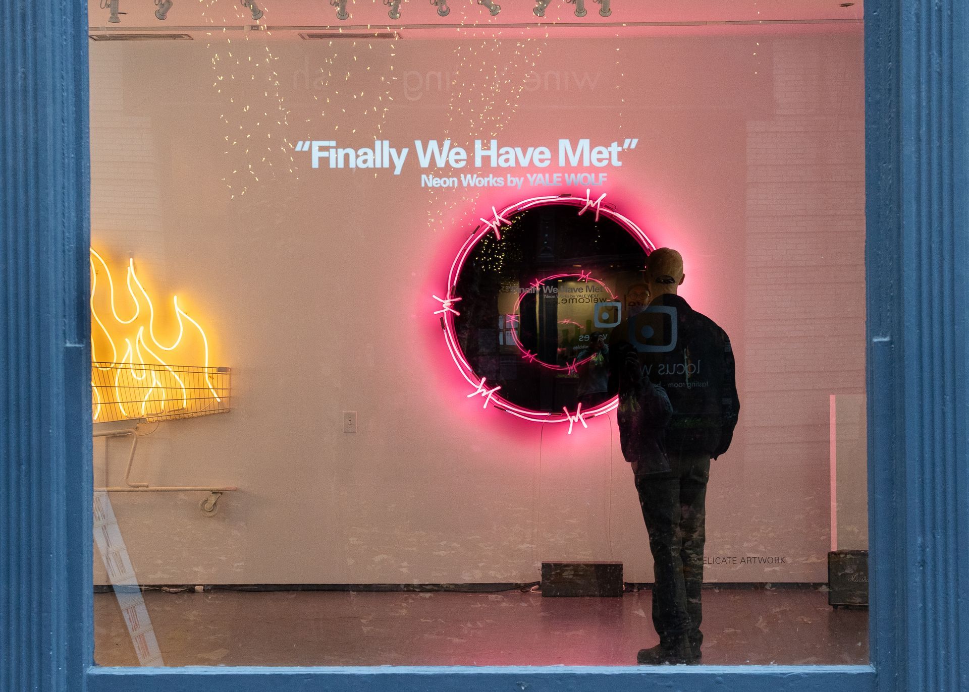 Man viewing neon art in a gallery window. A circular, pink-lit design and a yellow flame are visible; art by Yale Wolf. 