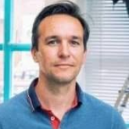 Een man met bruin haar, gekleed in een blauw poloshirt, kijkt in de camera.