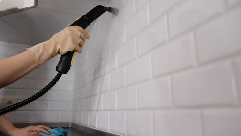 Hand holding a steam cleaner against a white tiled wall in a kitchen