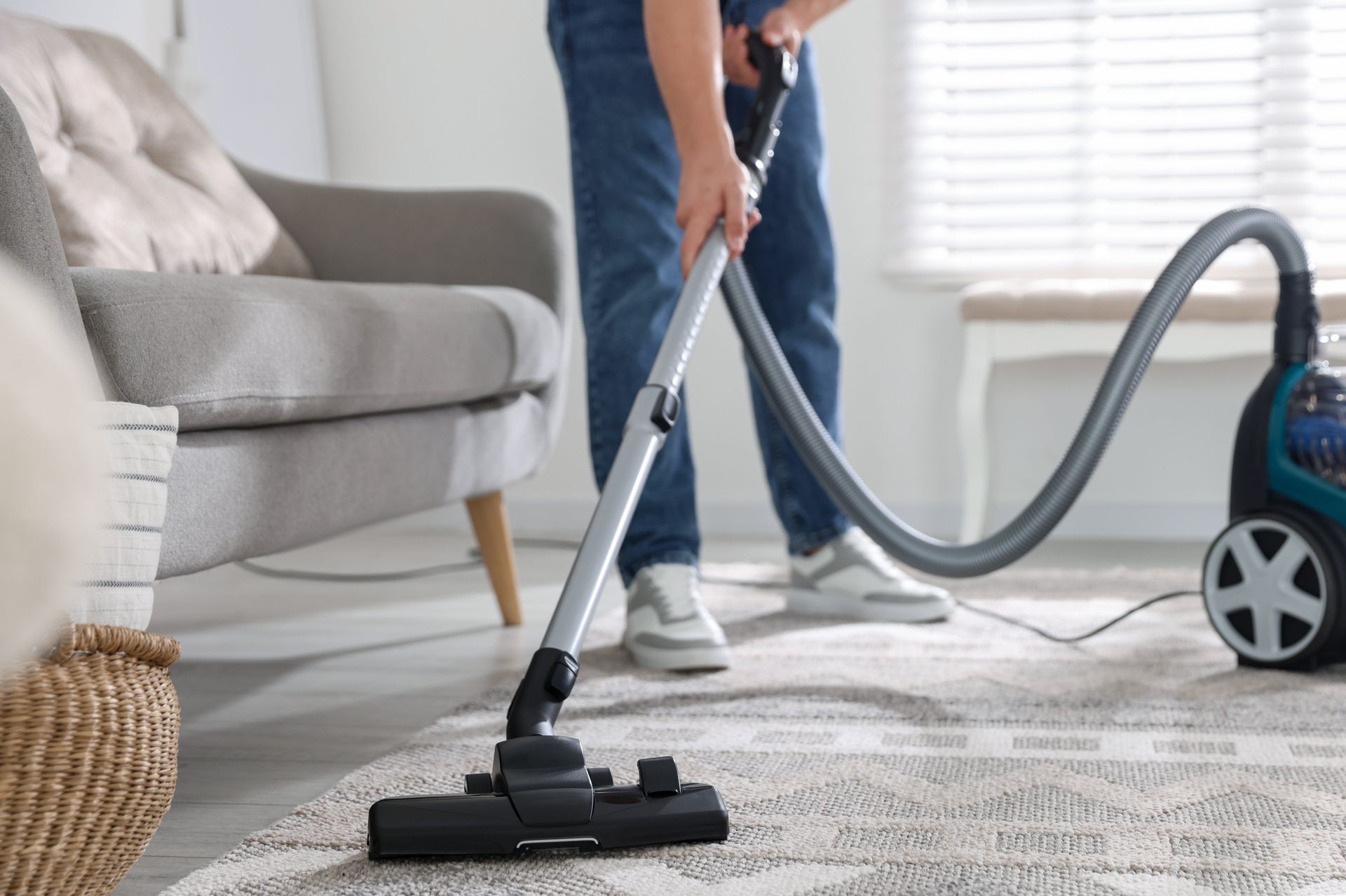 Person vacuuming a gray rug in a bright living room with a canister vacuum nearby