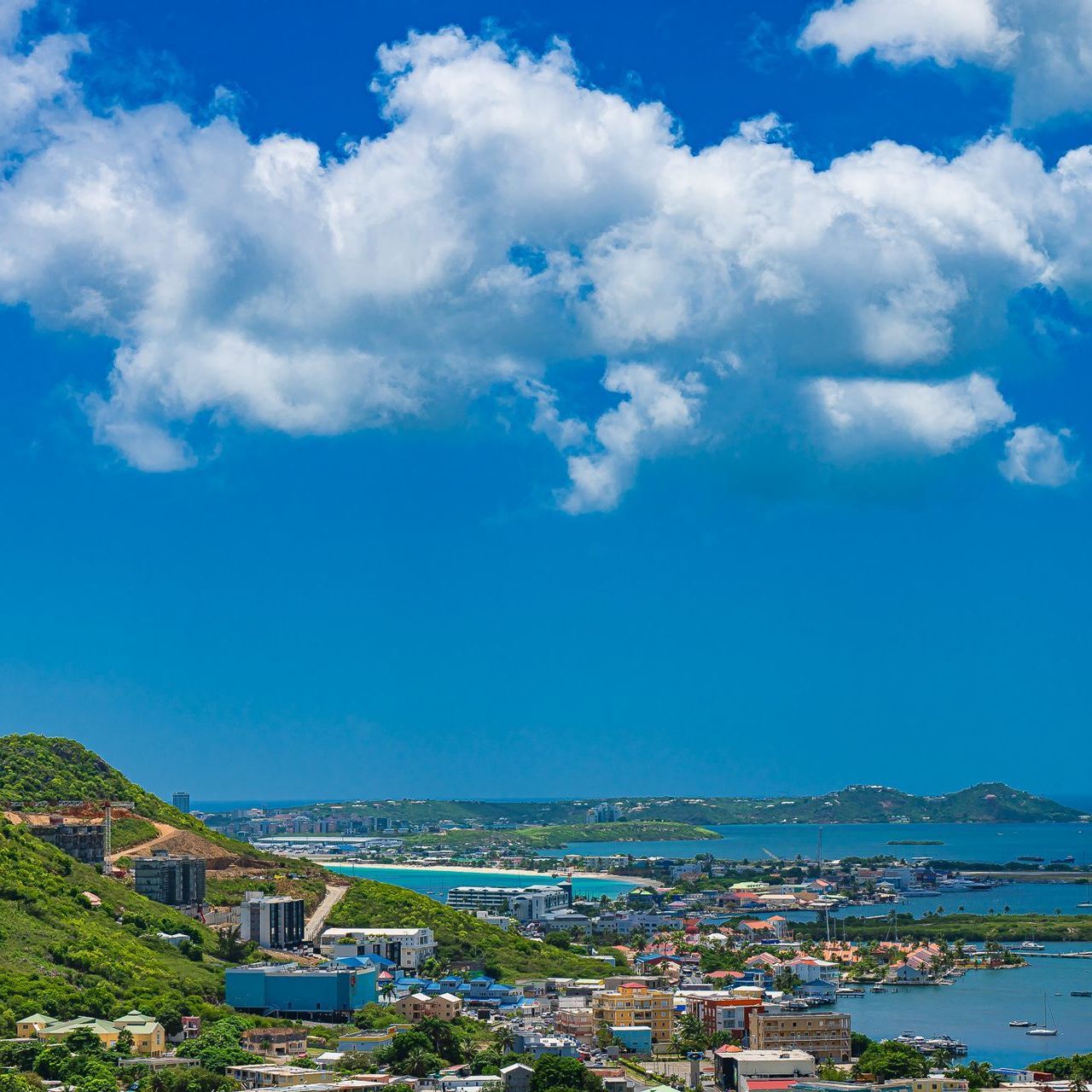 A sunny, vibrant coastal city with green hills, buildings, and a blue ocean under a cloudy sky.