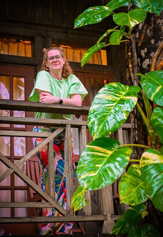 Woman in a light green top and colorful skirt smiles on a wooden balcony in Jamaica with lush green plants.