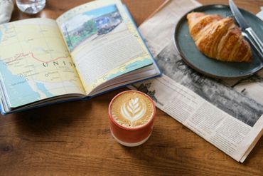 Latte with art, croissant on a plate, travel book, and newspaper on a wooden table.