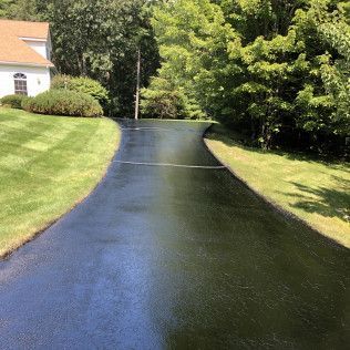 A black driveway leading to a house surrounded by trees and grass.