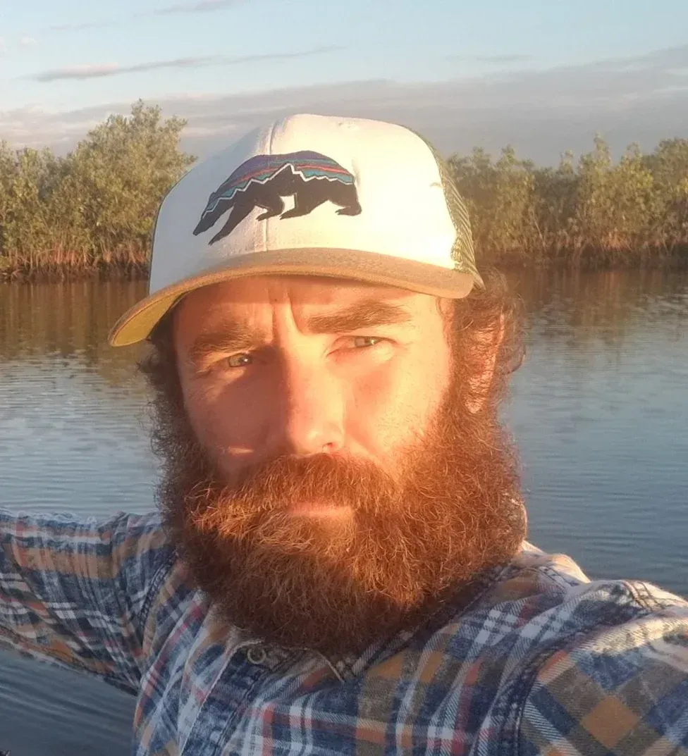 Bearded man in a plaid shirt and white cap taking a selfie on a calm river at sunset.