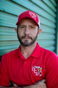 Man in red polo and cap, arms crossed, standing before a green corrugated door