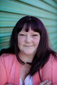 Smiling person with long dark hair wearing a pink cardigan against a teal striped background
