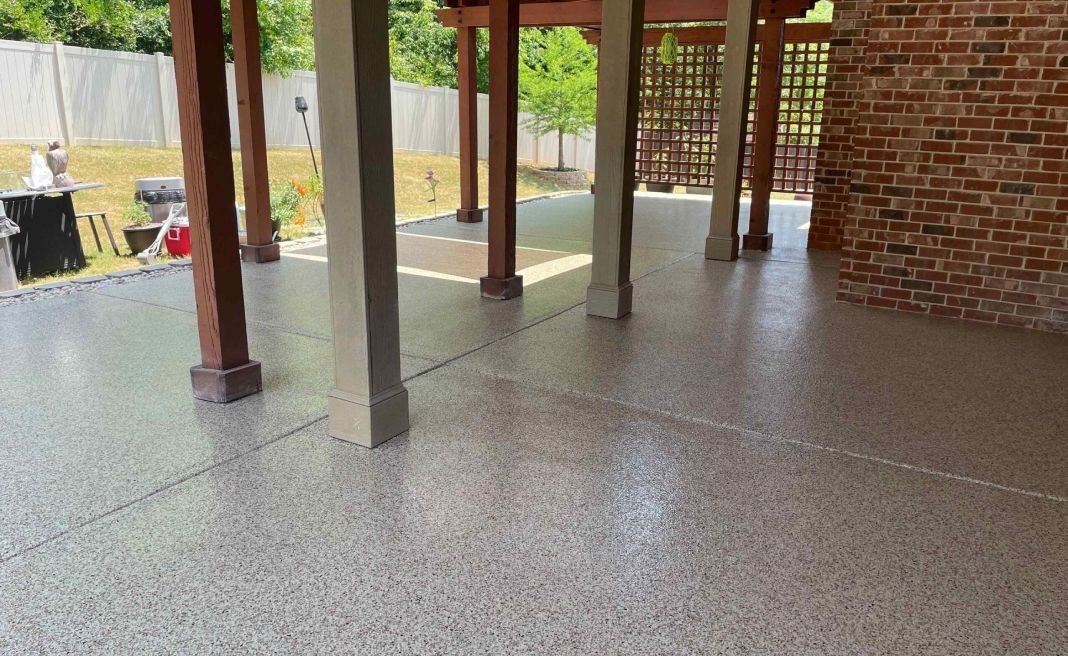 Covered brick patio with concrete columns and a speckled floor, opening to a yard and garden beyond