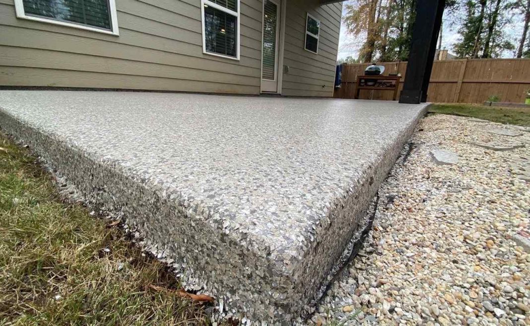 Freshly poured concrete patio beside a house, bordered by gravel and a wooden fence.
