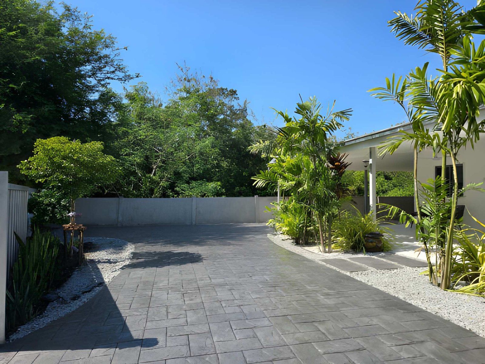 Sunlit paved driveway beside tropical plants and a white house under a clear blue sky