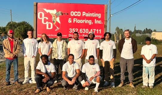 Group of workers posing in front of a red OCD Painting & Flooring Inc. sign outdoors