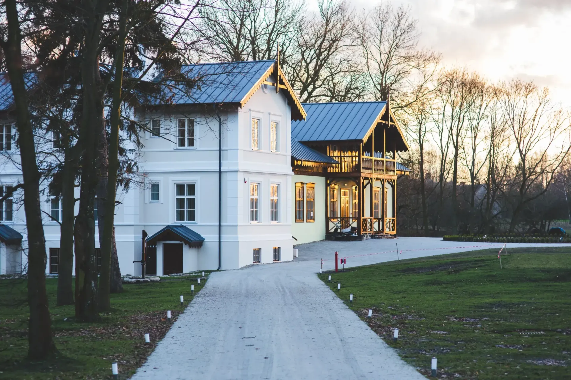 White farmhouse with a lit porch at dusk, set beside a gravel driveway and trees