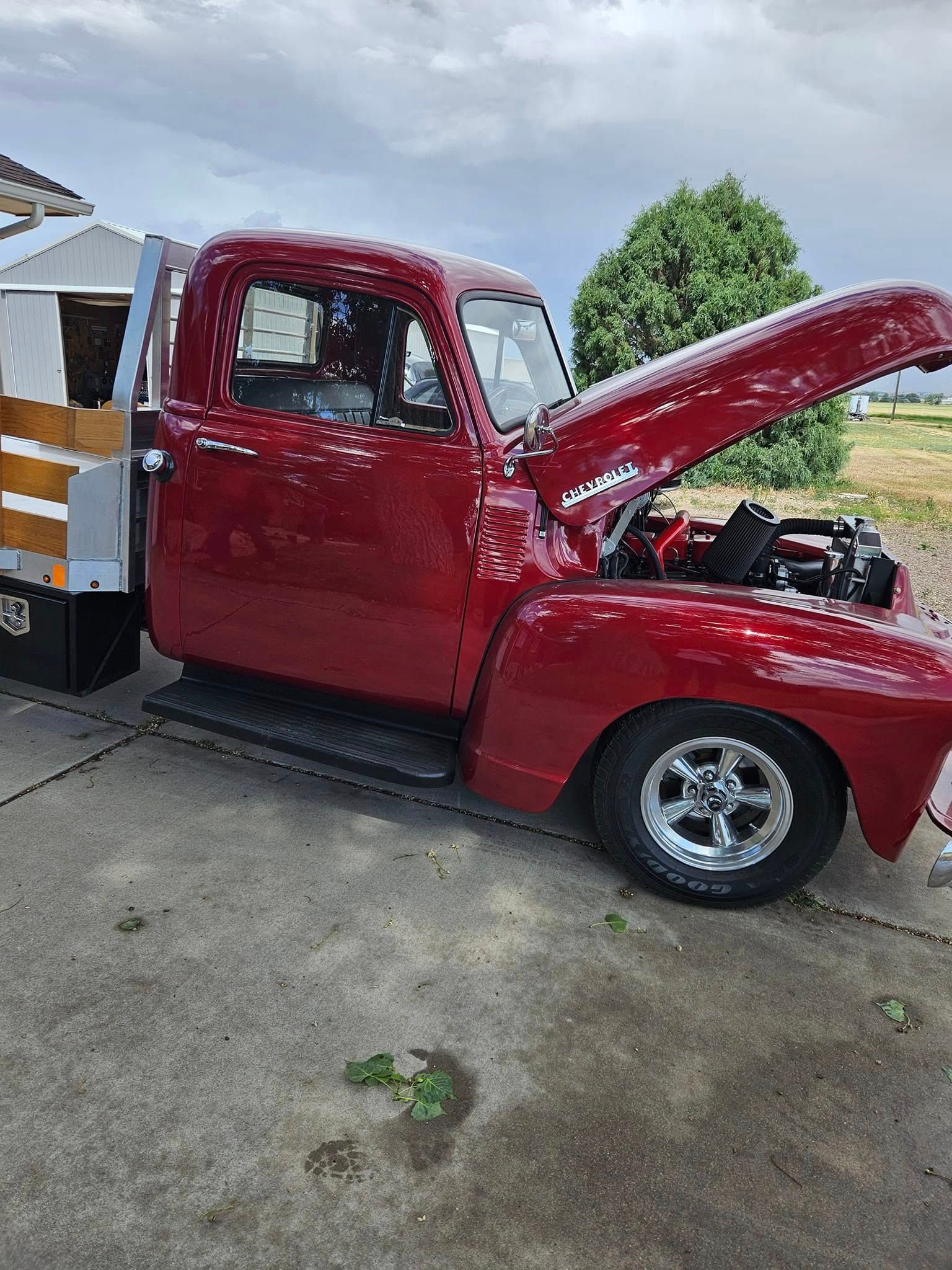 A shiny red vintage pickup truck with its hood open, parked on a concrete driveway under a cloudy sky.