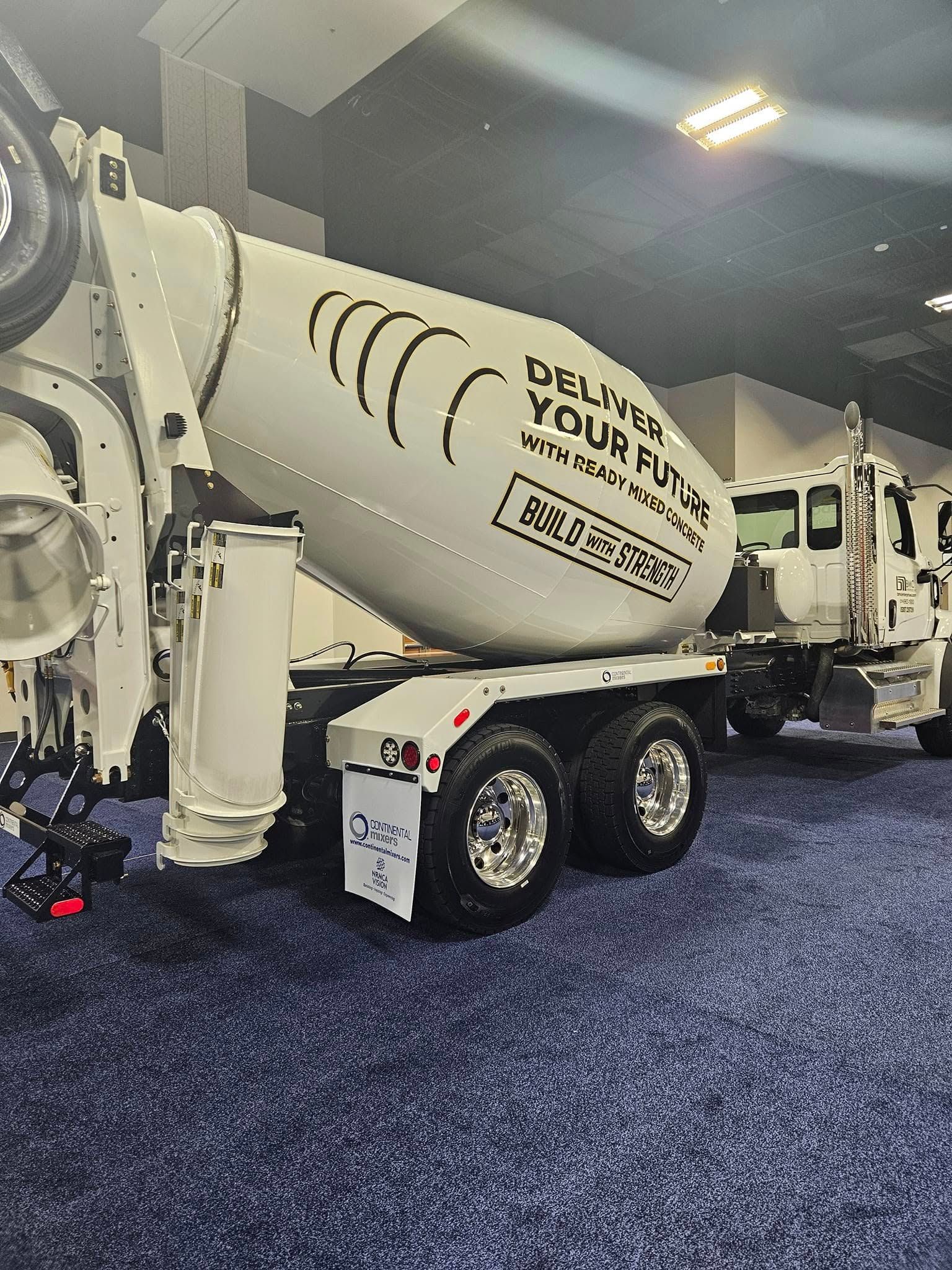 A white concrete mixer truck with branded signage parked indoors on a dark blue carpeted floor under bright lights.